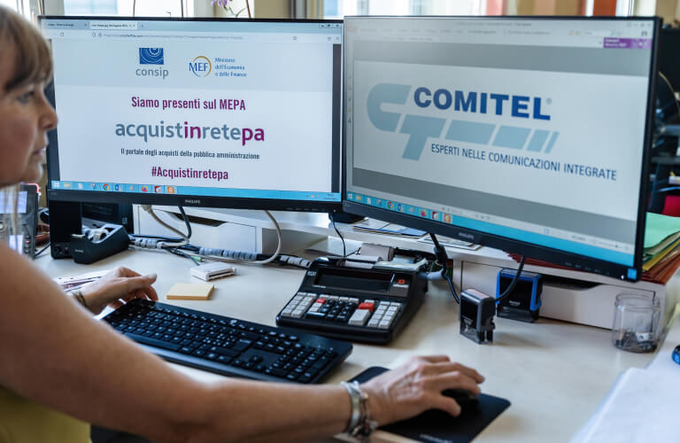 Foto che ritrae una persona al computer con due schermi che mostrano la presenza di Comitel sul MEPA